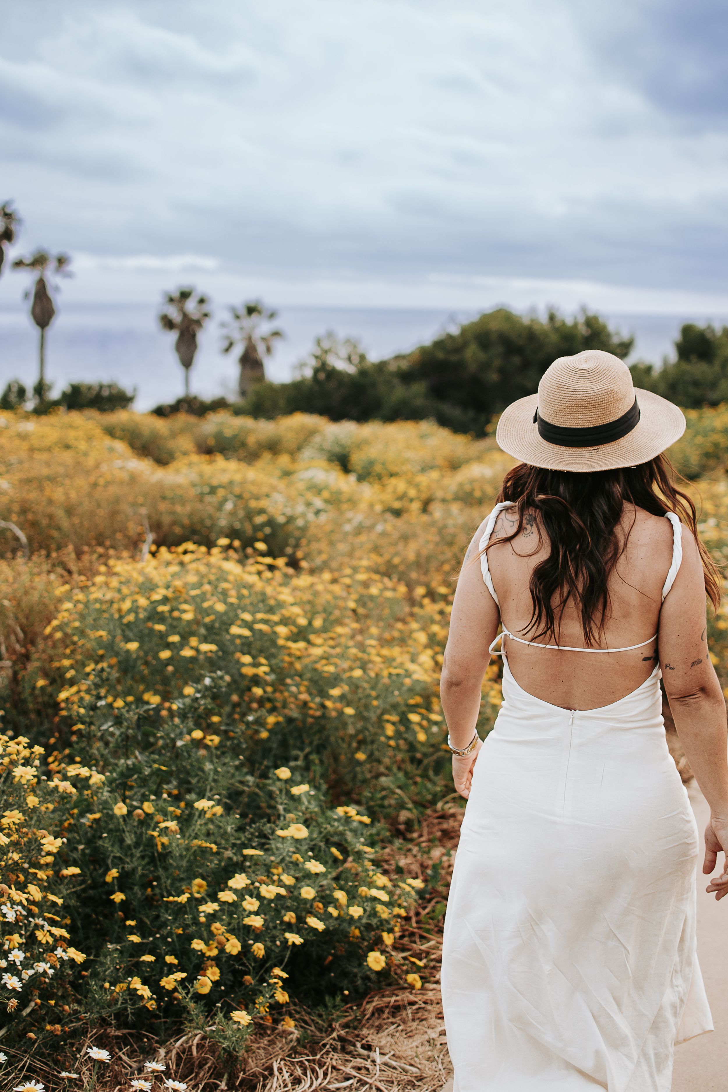 Woman in white dress walks through field of yellow flowers with palm trees in background.