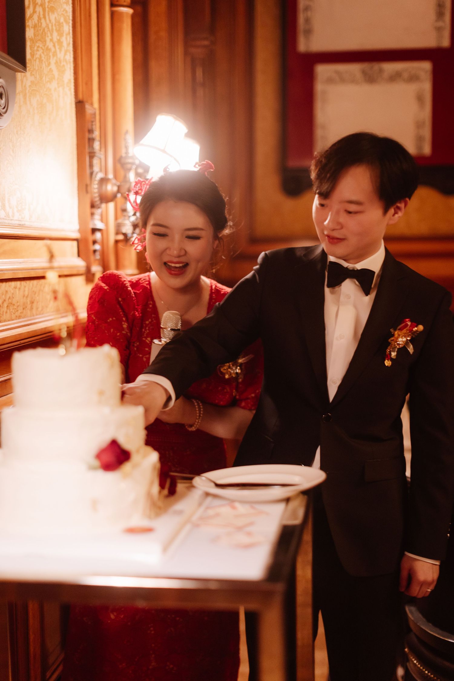 Couple in formal attire cutting their wedding cake in an elegant venue with warm lighting.