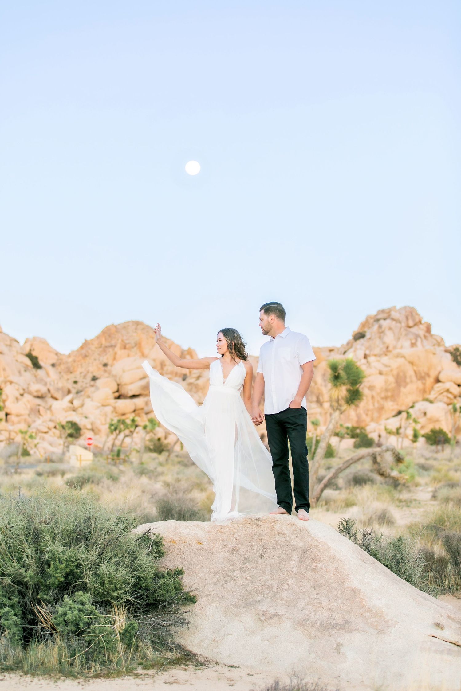 A woman throws her dress in the air as she stands on a rock with her fiance in Joshua Tree with the moon behind them.
