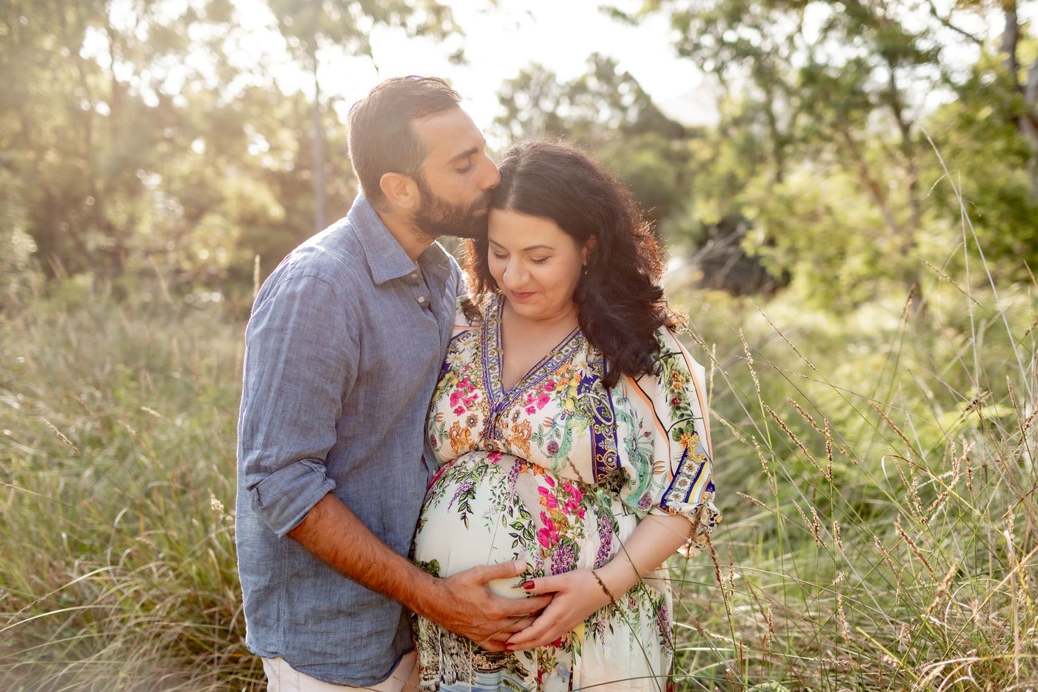 Maternity photo session in a sunlit field with a floral dress and denim shirt during golden hour.