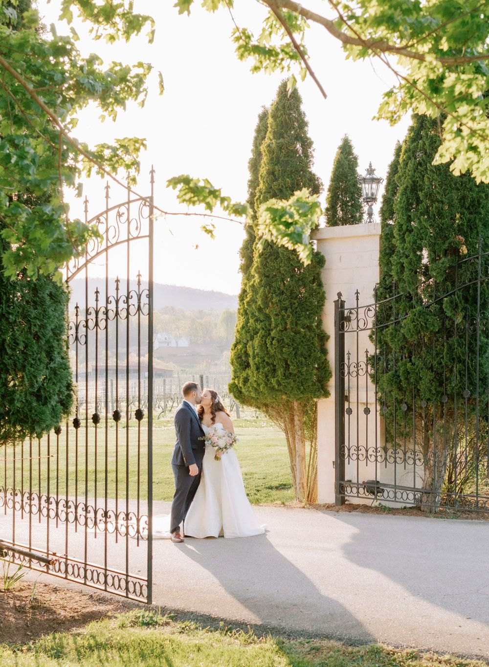 Wedding couple shares an intimate moment by ornate iron gates with cypress trees in golden sunlight.