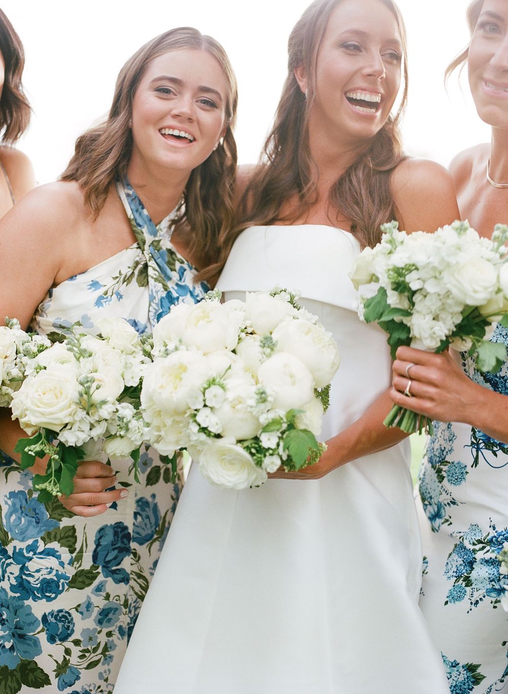 Bridesmaids in floral blue dresses hold white bouquets while celebrating with the bride.