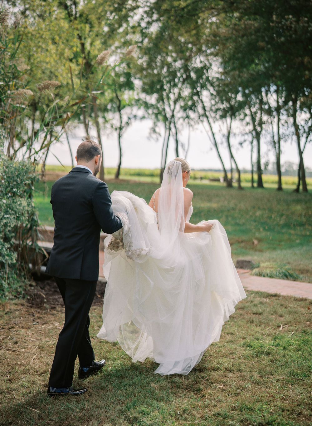 Groom helps bride with flowing dress during outdoor wedding photoshoot in nature.