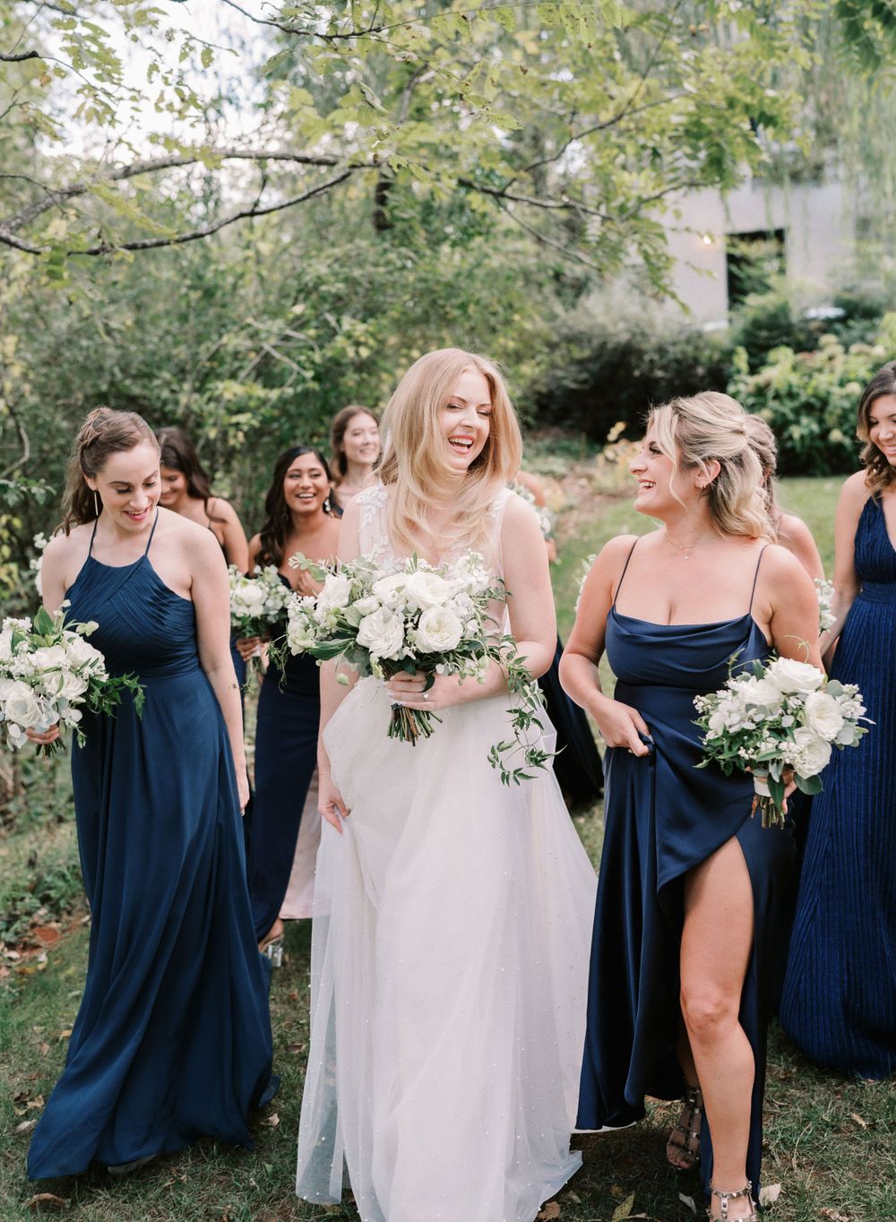 Bridesmaids in navy blue dresses holding white bouquets laugh together with the bride in a garden setting.
