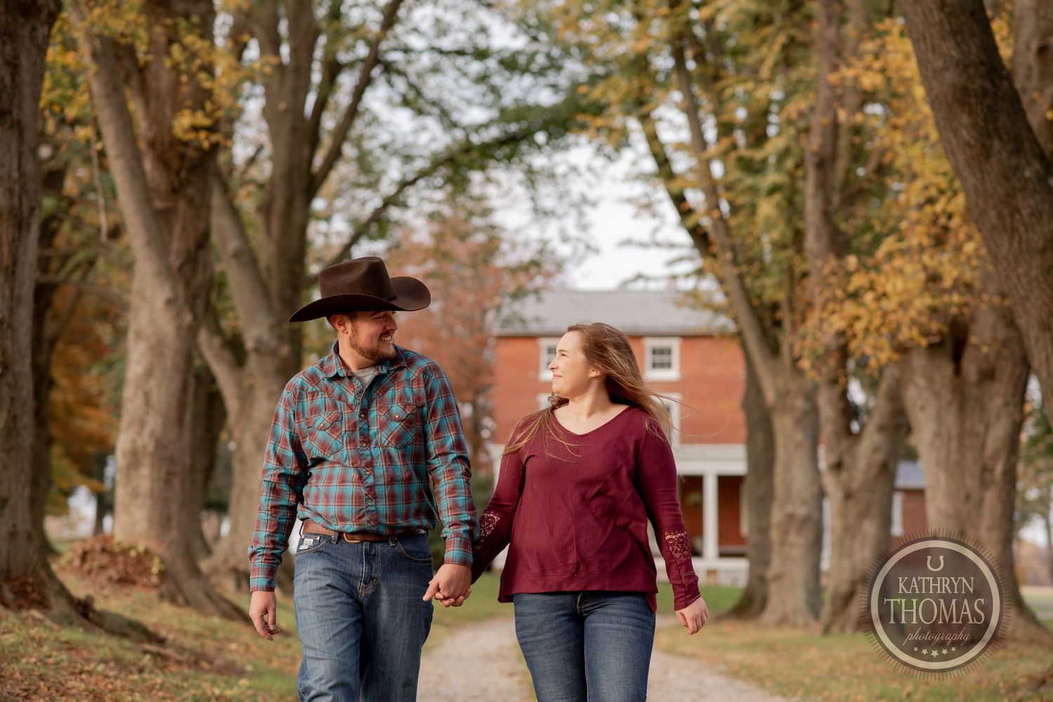 A couple walks along a tree-lined pathway with fall foliage and a red brick house behind them.