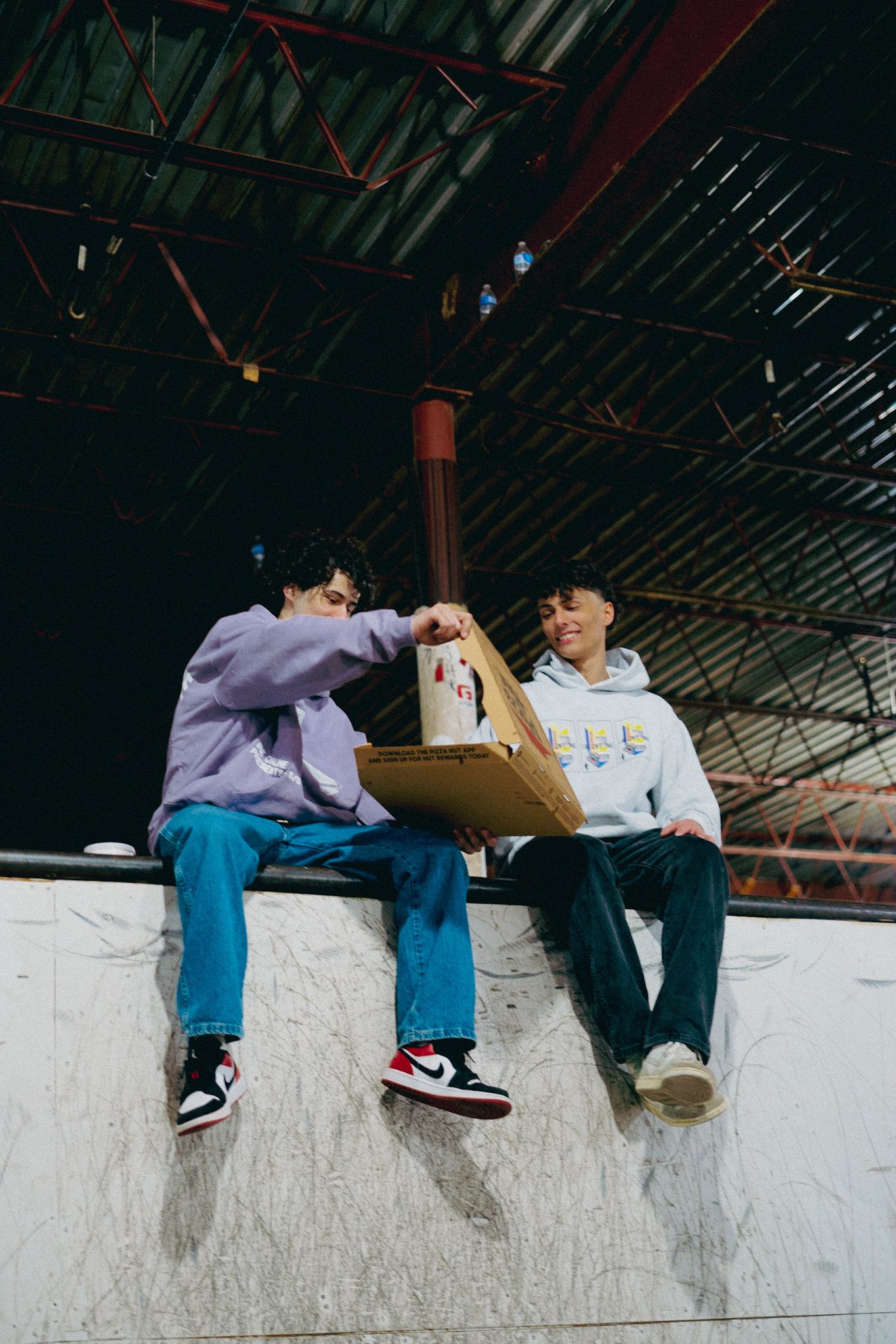 Two people sit casually on a white wall while sharing food or snacks.