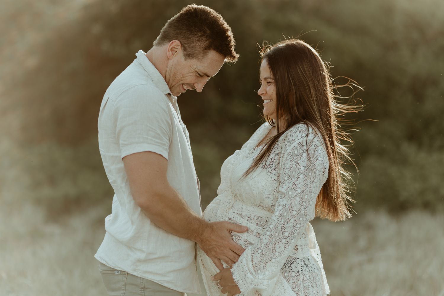 Expectant couple shares intimate moment in sunlit field at golden hour.
