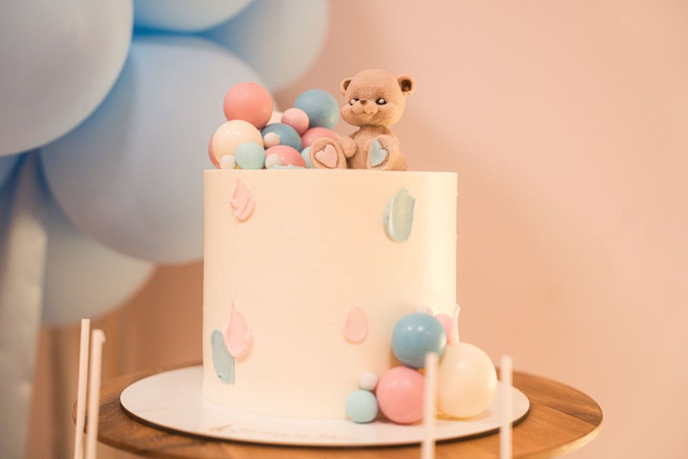 White cake decorated with pastel blue and pink accents and a teddy bear topper for a baby shower.