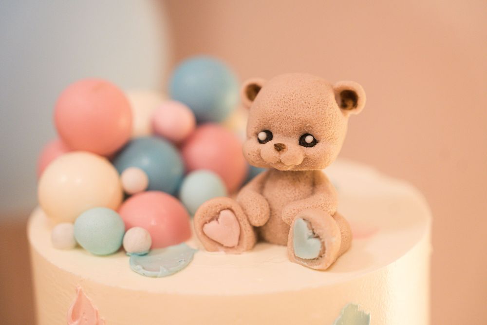 Elegant white cake with pastel decorations and a plush teddy bear on display.