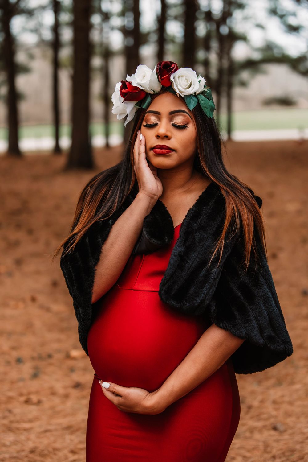 Maternity photo shoot featuring a red dress, black fur shawl, and floral crown in a forest setting.