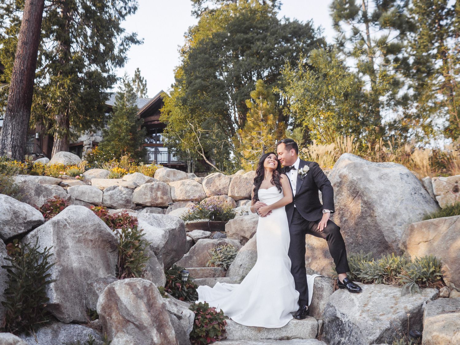 A couple in wedding attire pose together on natural stone steps surrounded by trees and landscaping at sunset.