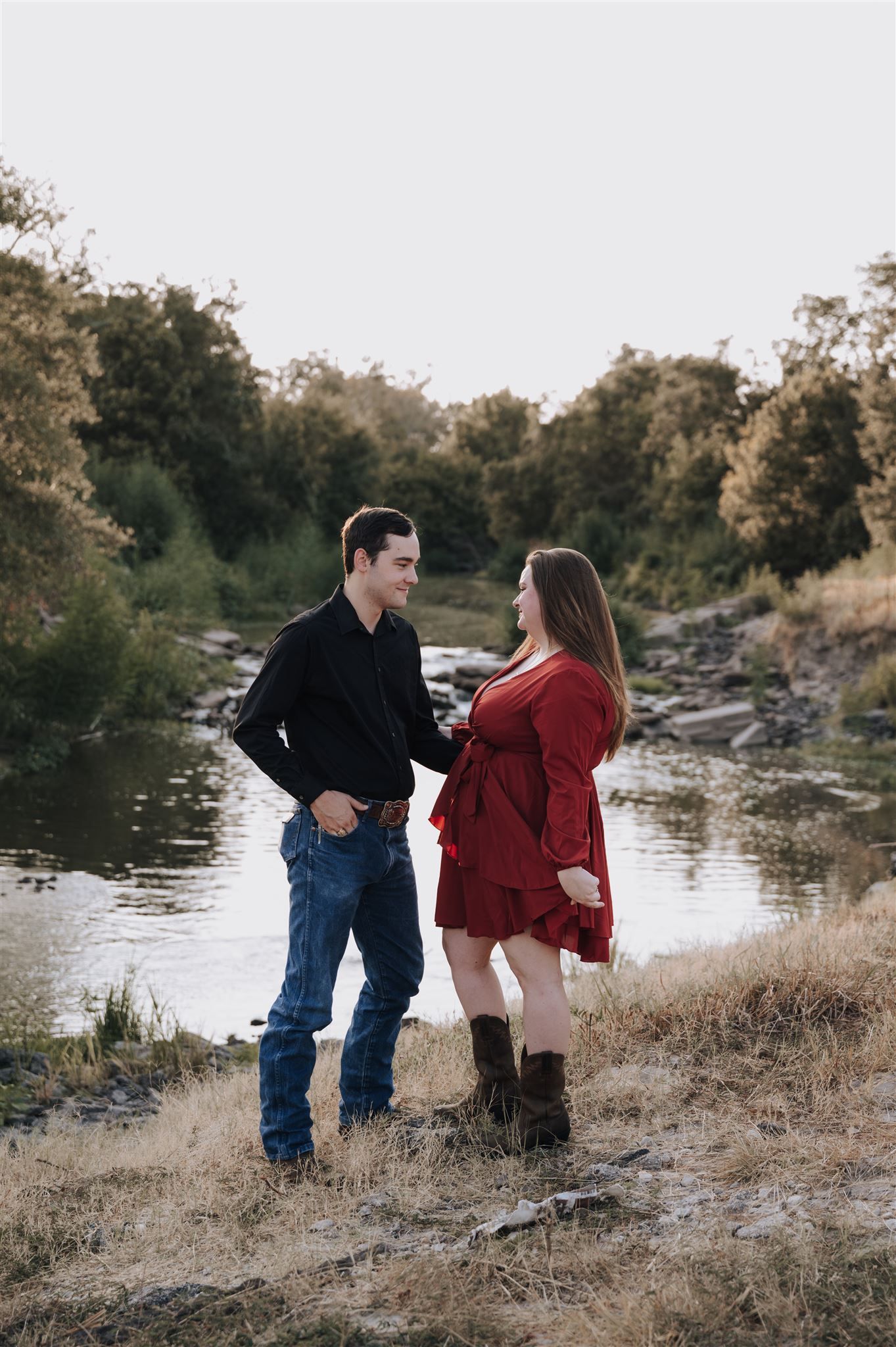 A couple stands together by a river during a sunset photoshoot.