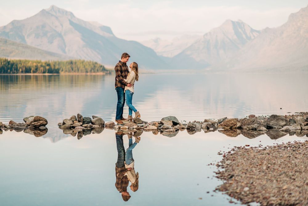 Couple embraces on rocky shoreline with mountain lake reflections at sunset in scenic wilderness landscape.