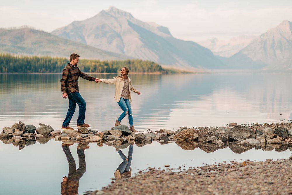 Two people walking on rocky shore with mountains and lake reflections in background.