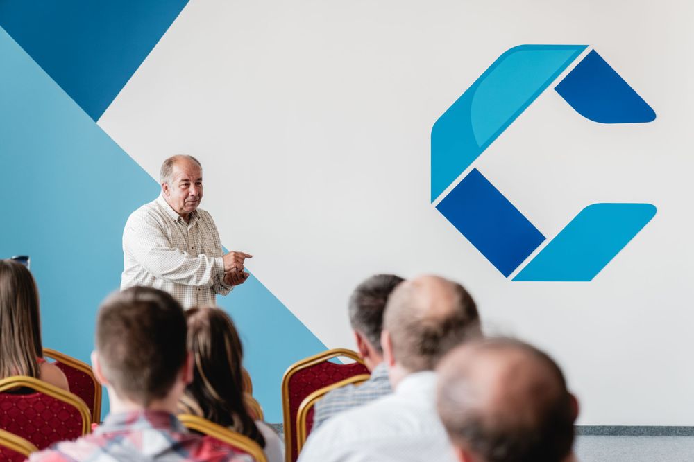 Professional speaker addresses seated audience with blue corporate branding on white wall.