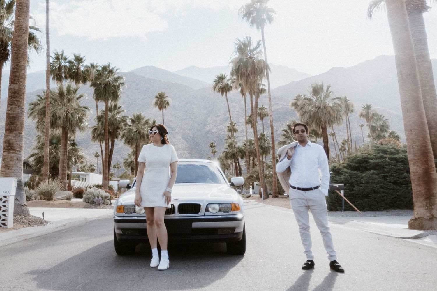 Retro-styled couple poses with vintage BMW car on palm tree-lined street.