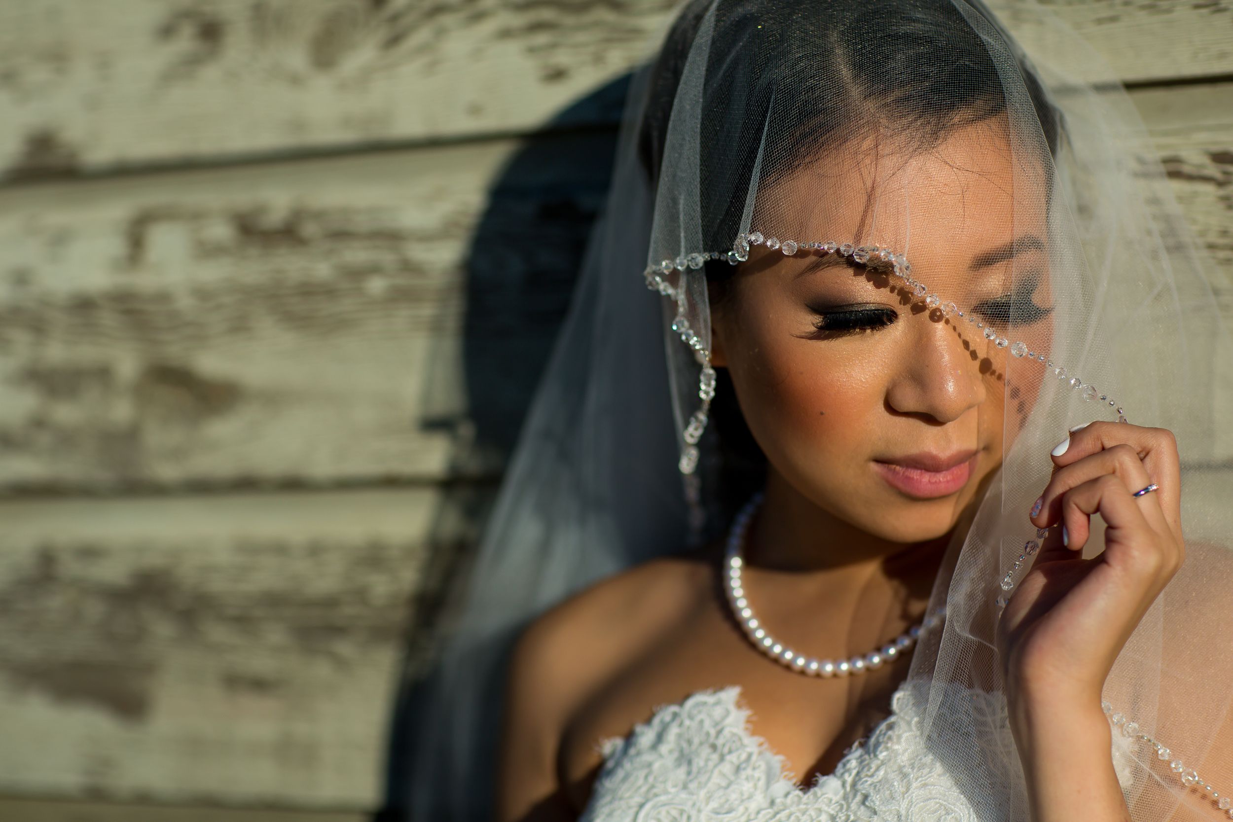 A bride in white lace wearing pearl jewelry and a sheer veil poses against a rustic wooden backdrop.