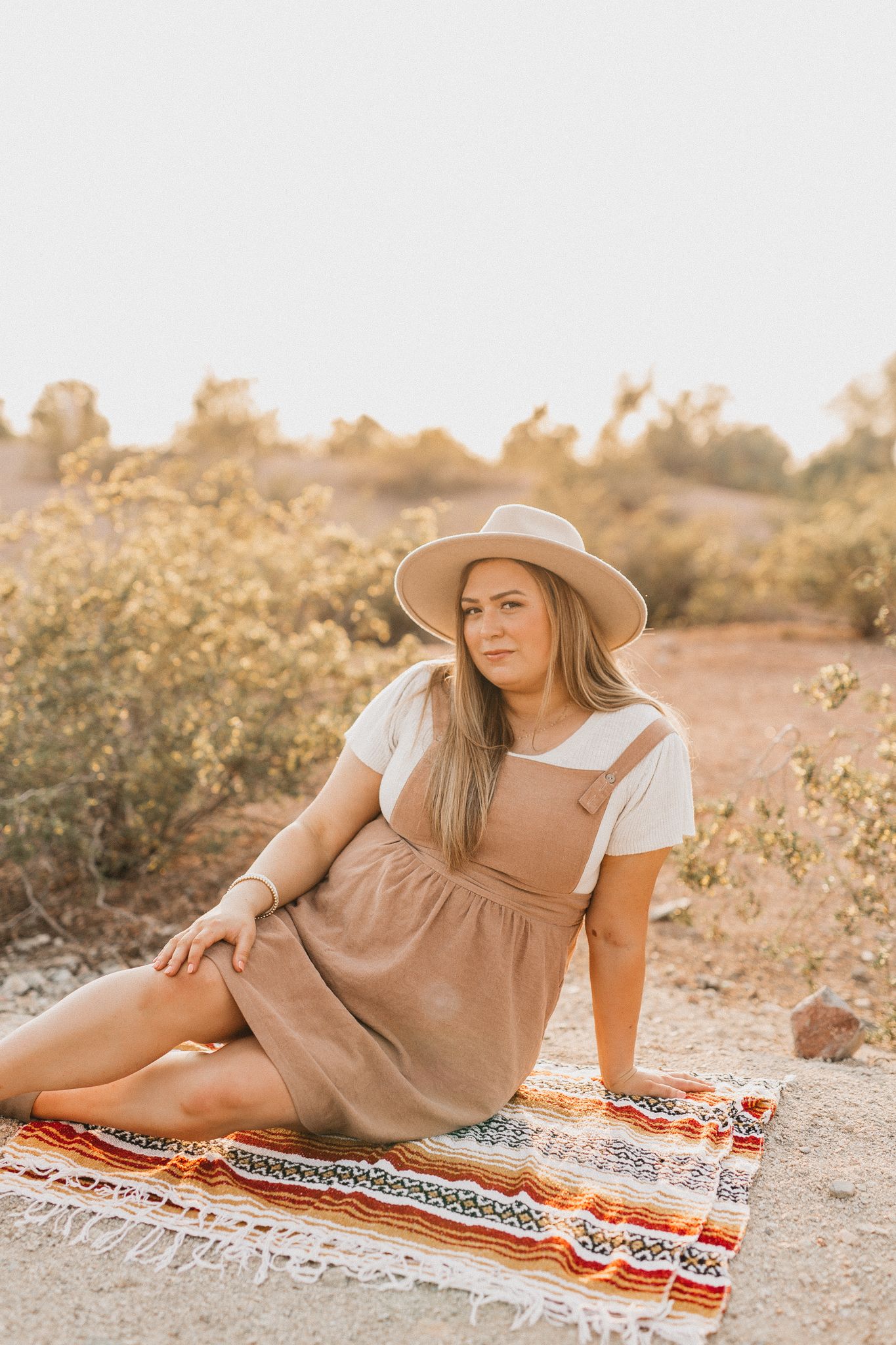 Woman in brown dress sits on blanket in desert setting wearing wide brim hat.
