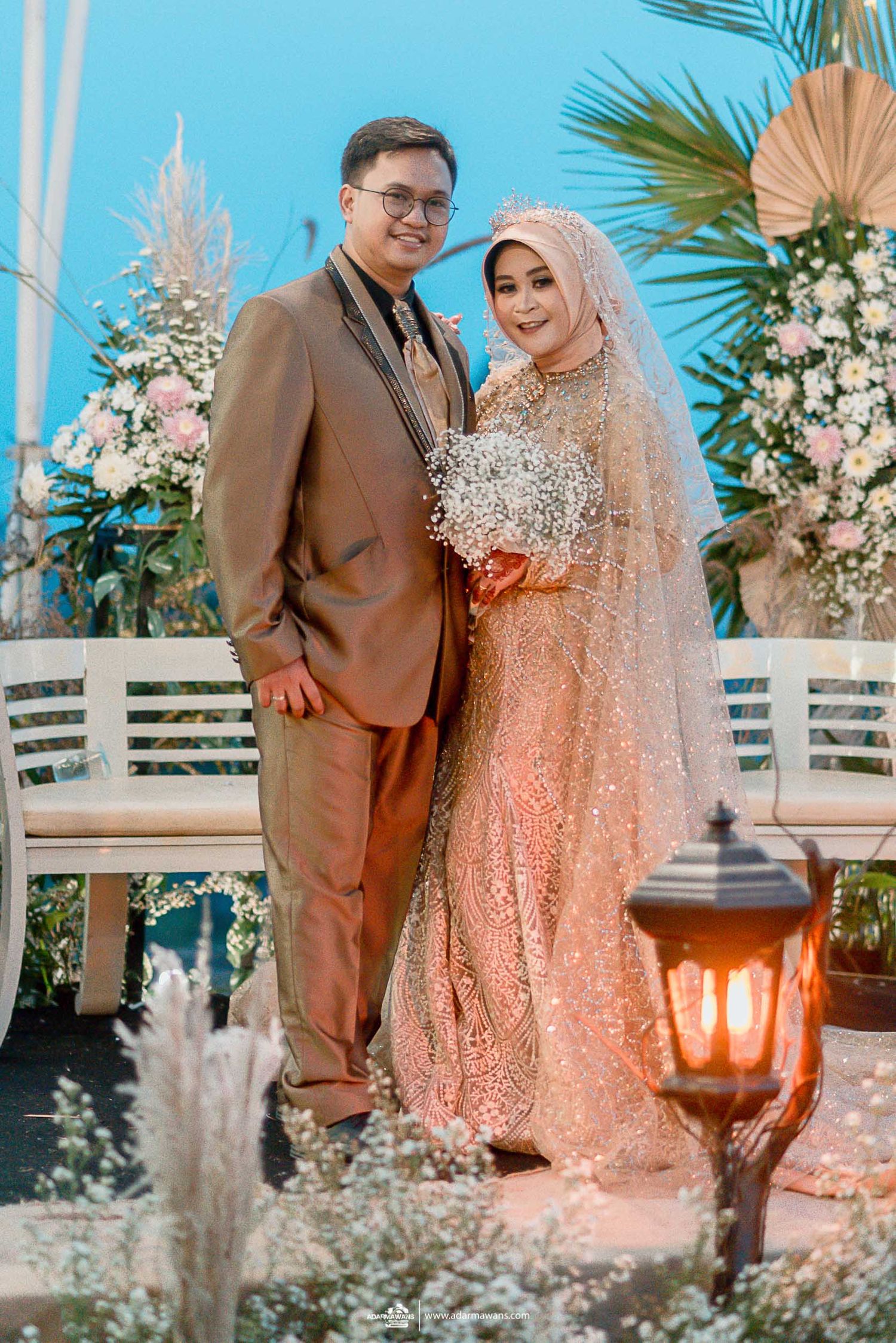 Wedding couple in traditional attire posing against floral backdrop with palm leaves.