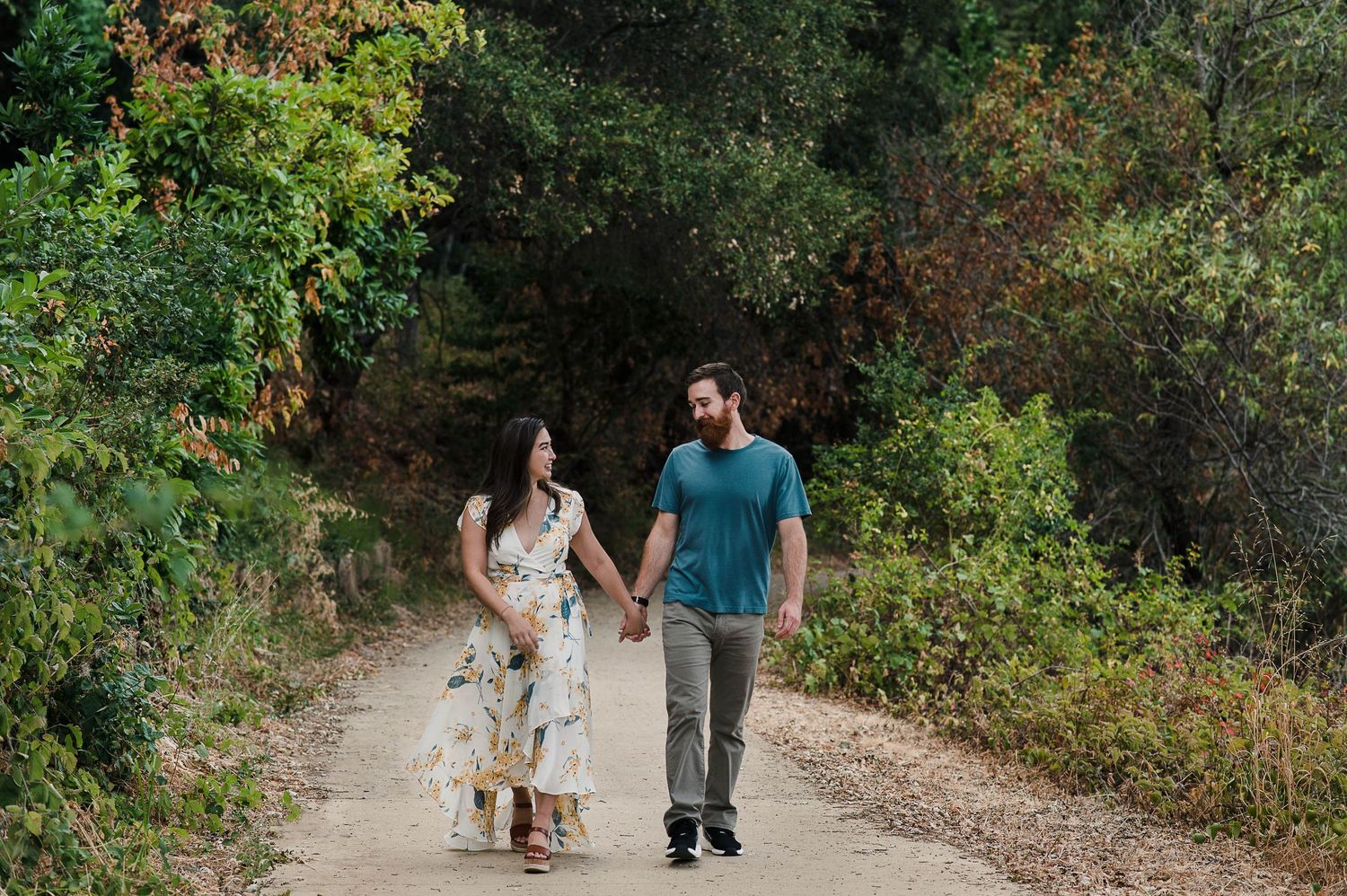 A couple walks hand in hand down a sunlit nature trail surrounded by autumn foliage.