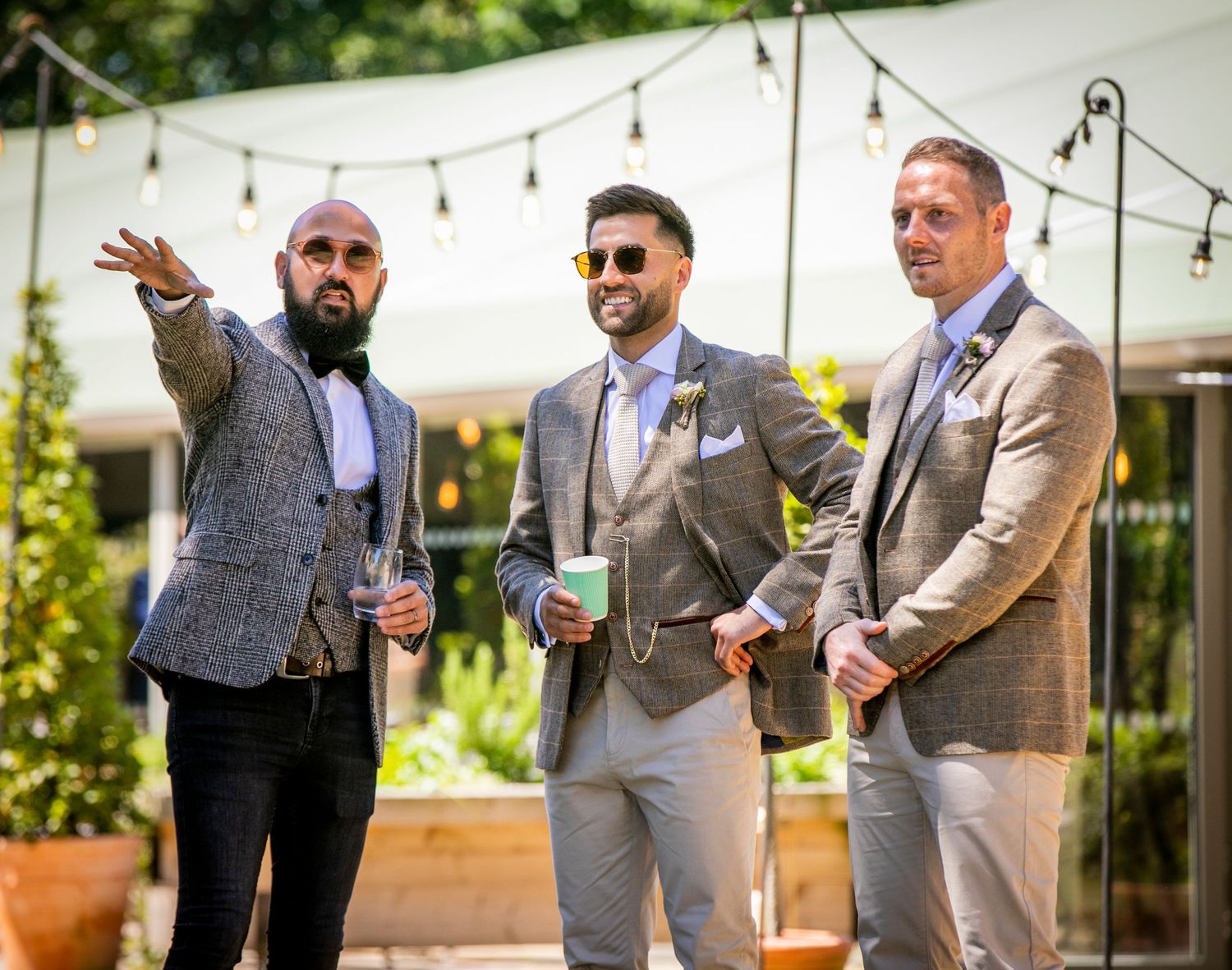 Three groomsmen in suits chat and gesture during an outdoor wedding reception.