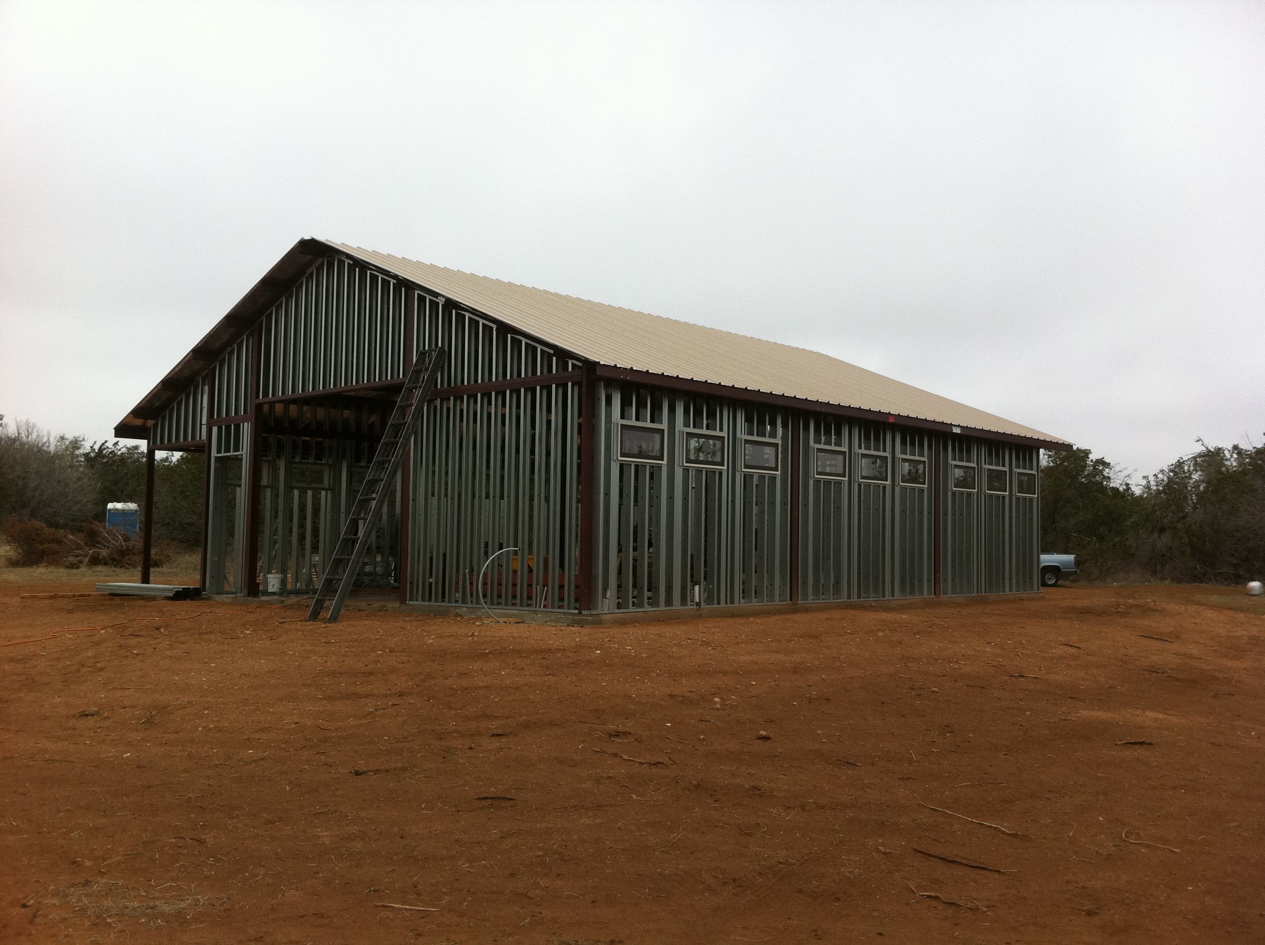 A panoramic view of a long green barn structure on a brown dirt field under an overcast sky.