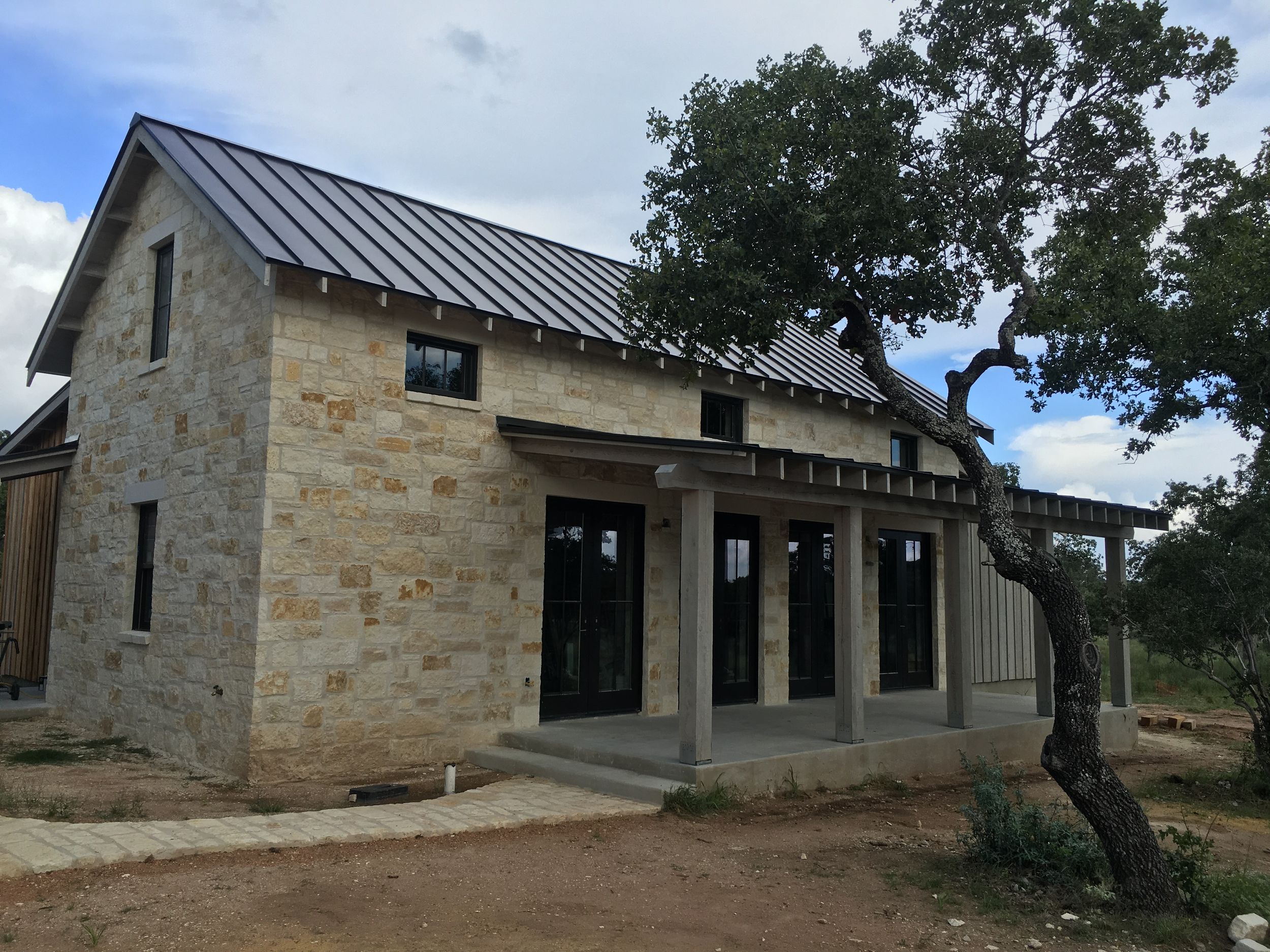 Modern farmhouse exterior features limestone walls, metal roof, and large windows set against a rustic Texas Hill Country landscape.