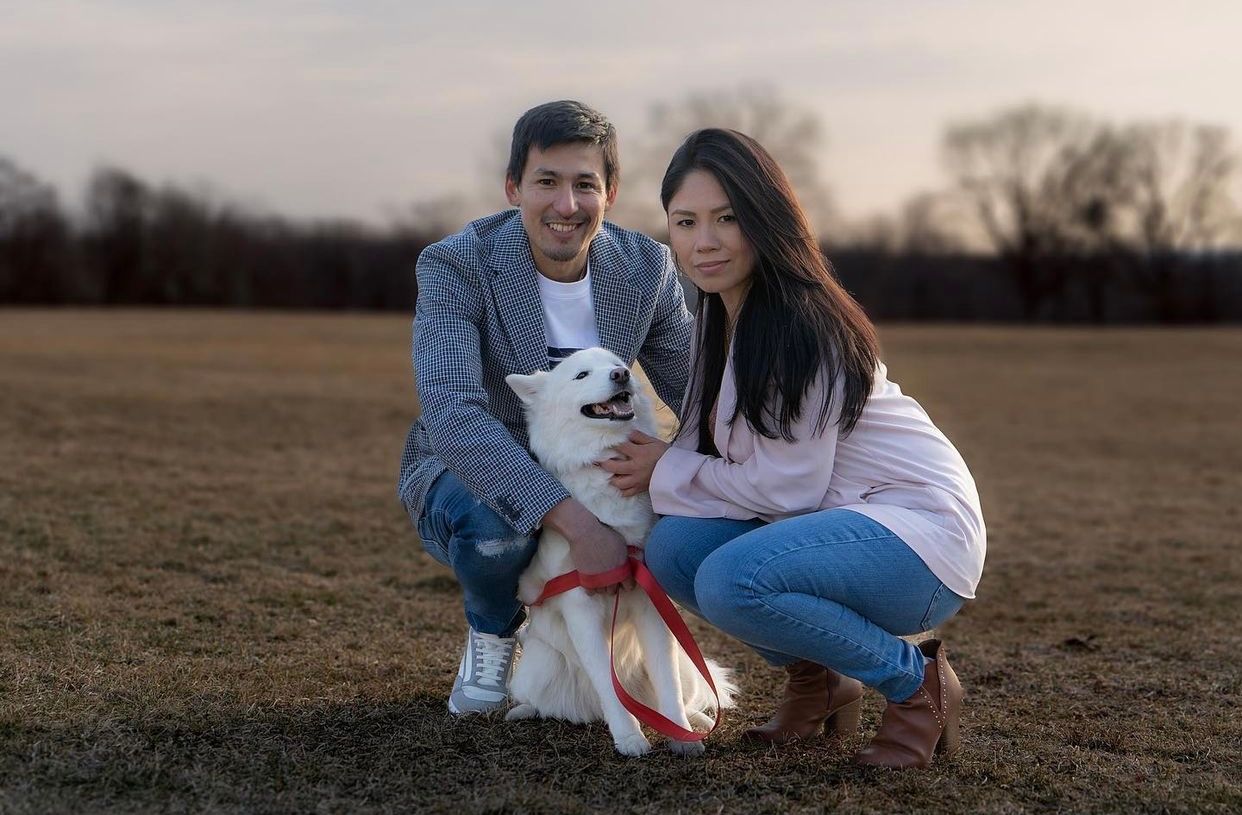 Couple posing with their white and brown dog in an open field at sunset.