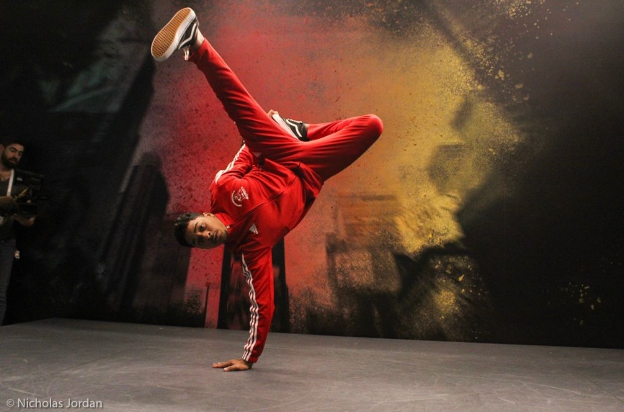 Breakdancer performing a one-handed freeze move against colorful backdrop.