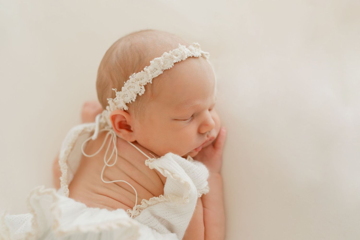 Sleeping newborn wearing delicate white lace headband and dress in soft neutral tones.
