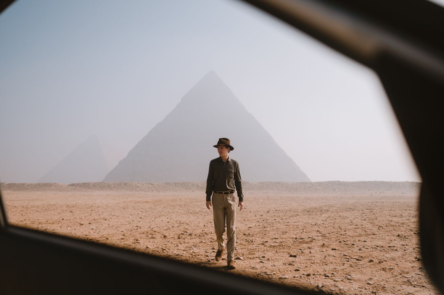 Silhouette of a person standing in front of Egyptian pyramids viewed through a car window.