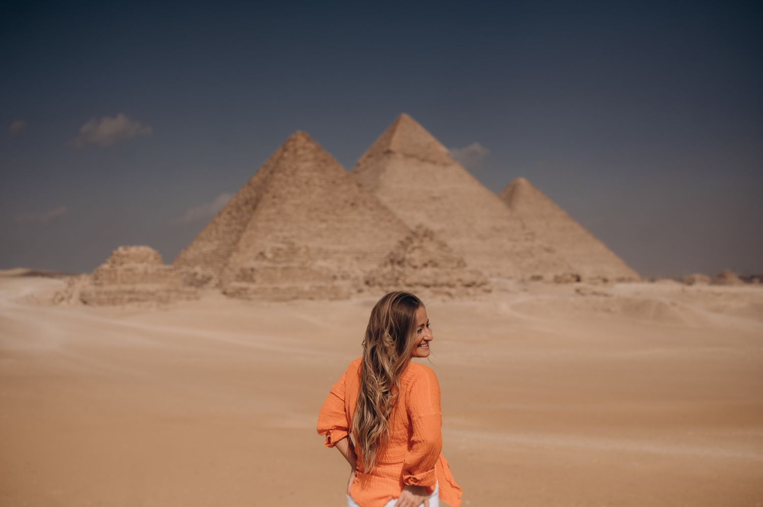 Woman in orange sweater looking at the majestic pyramids of Giza in the desert.