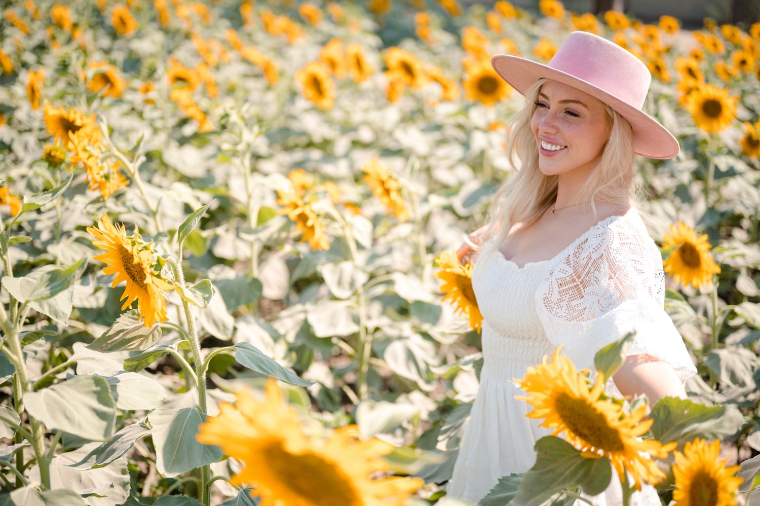 Woman in white dress and pink hat standing in a vibrant field of blooming sunflowers.