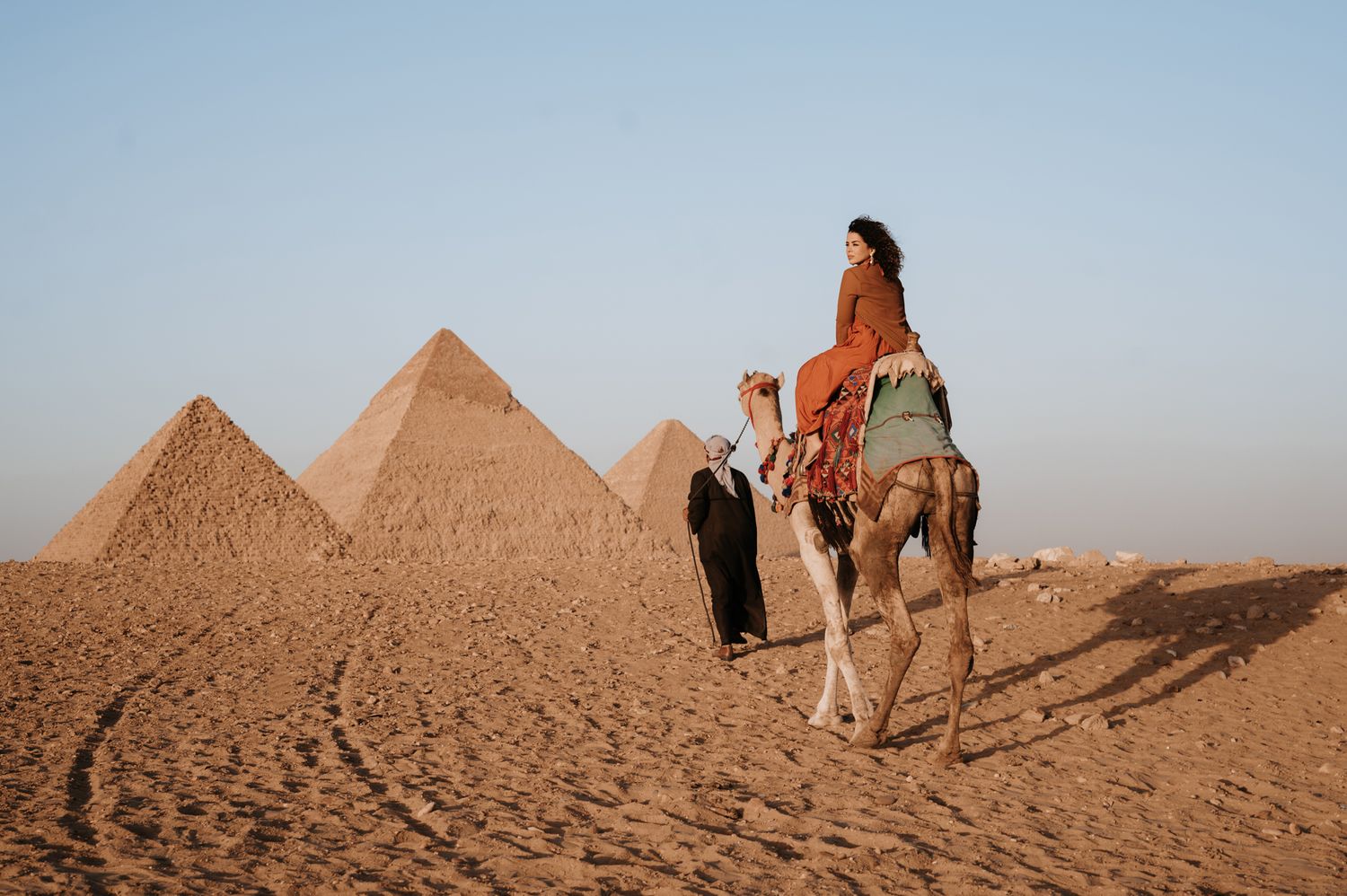 Desert travelers riding a camel with the iconic pyramids of Giza in the background.