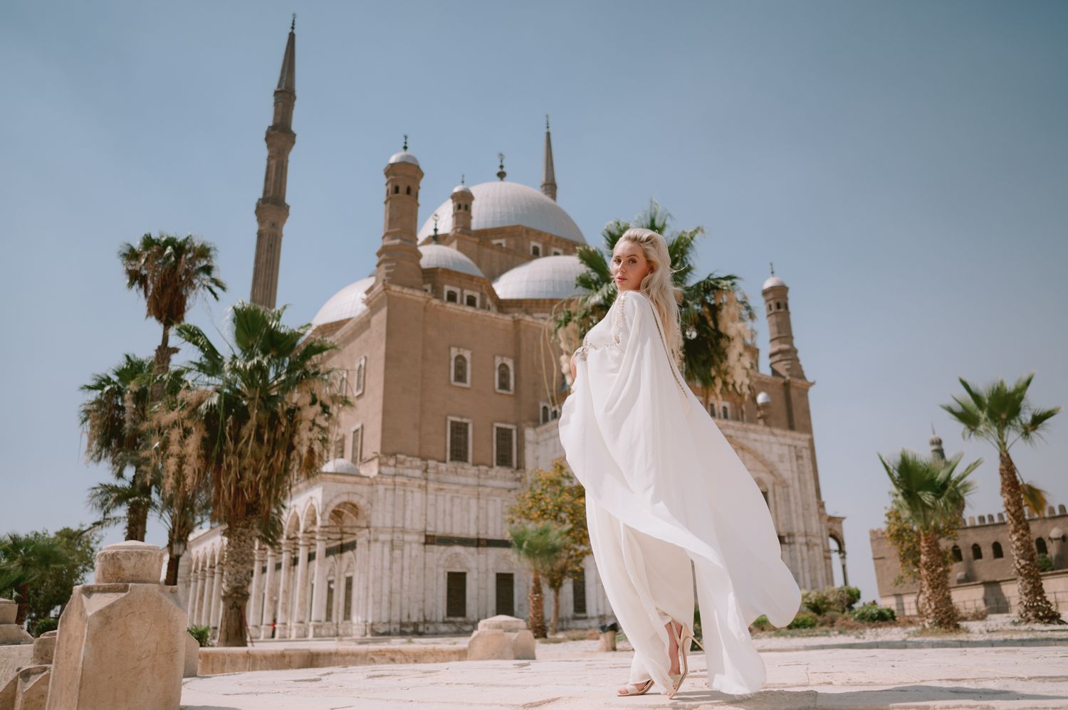 White flowing dress billowing in front of a majestic mosque with palm trees and minarets.