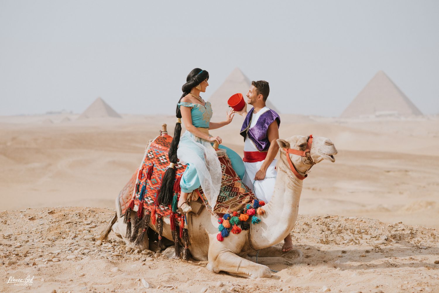 Tourists riding decorated camels with pyramids of Giza visible in the desert background.