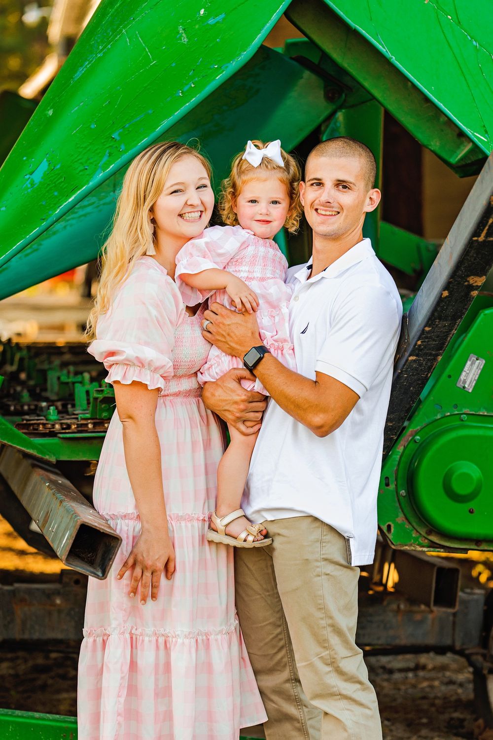 Family poses together near green tractor in pink and white outfits.