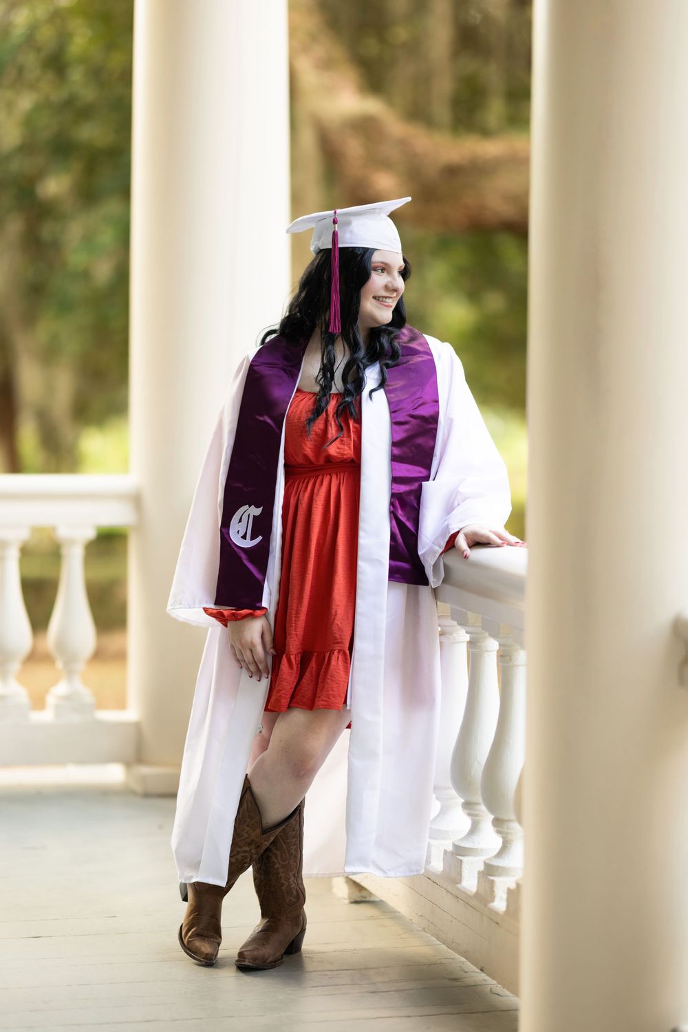 Graduate wearing red dress and cowboy boots poses on white columned porch.