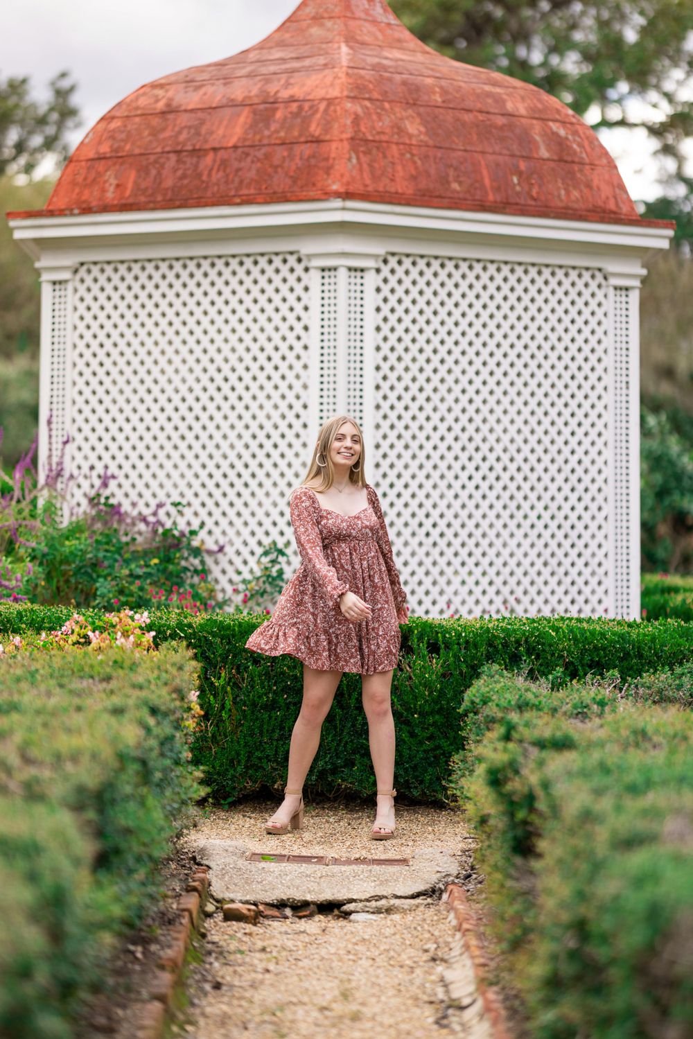 Young person in floral dress poses in garden setting with white lattice gazebo and copper dome.