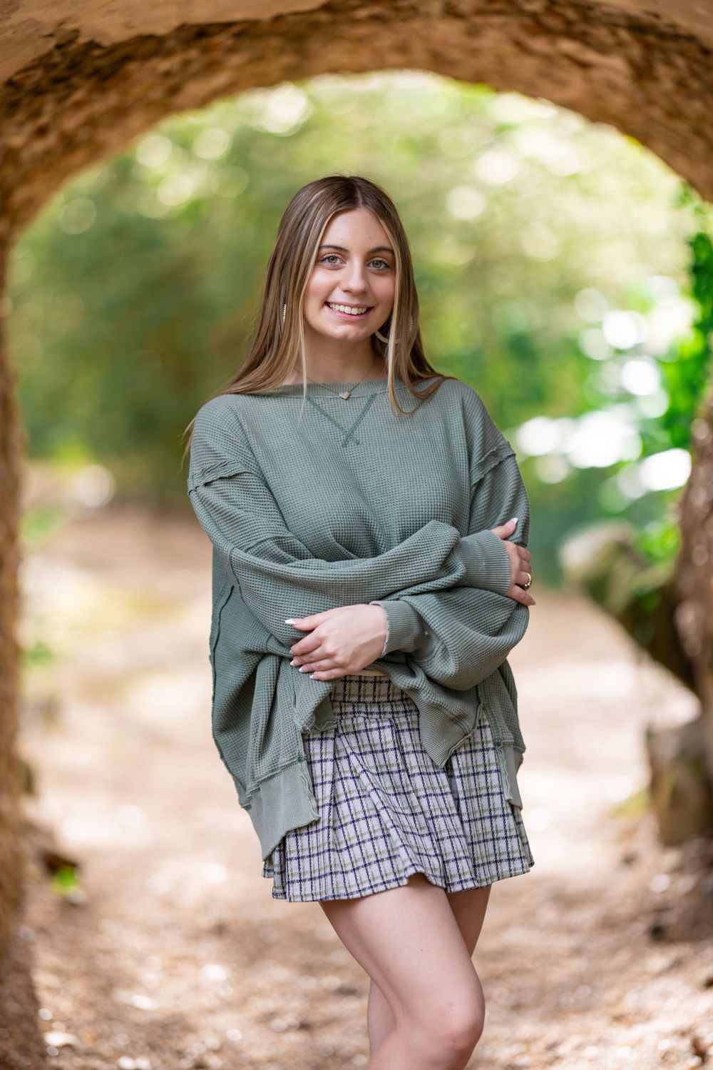 Person in green sweater and plaid skirt poses under stone archway with bokeh background.