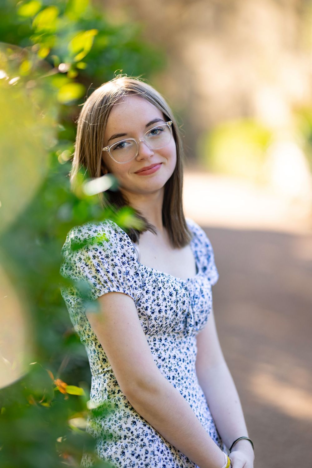 A person in a floral dress poses among green foliage in soft natural lighting.