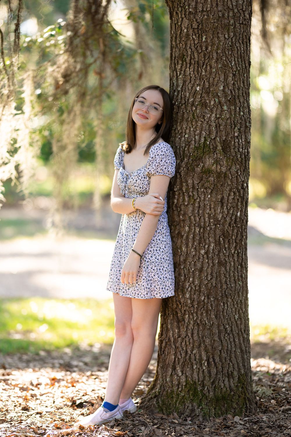Person in floral dress poses against large tree trunk in sunlit forest setting.