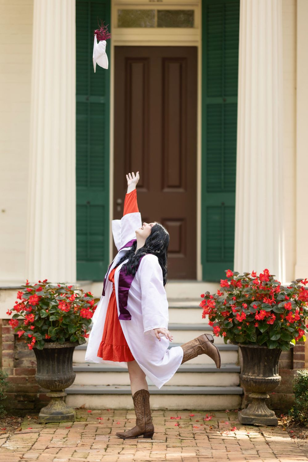 Graduate in white cap and gown jumping joyfully in front of historic doorway with red flowers.