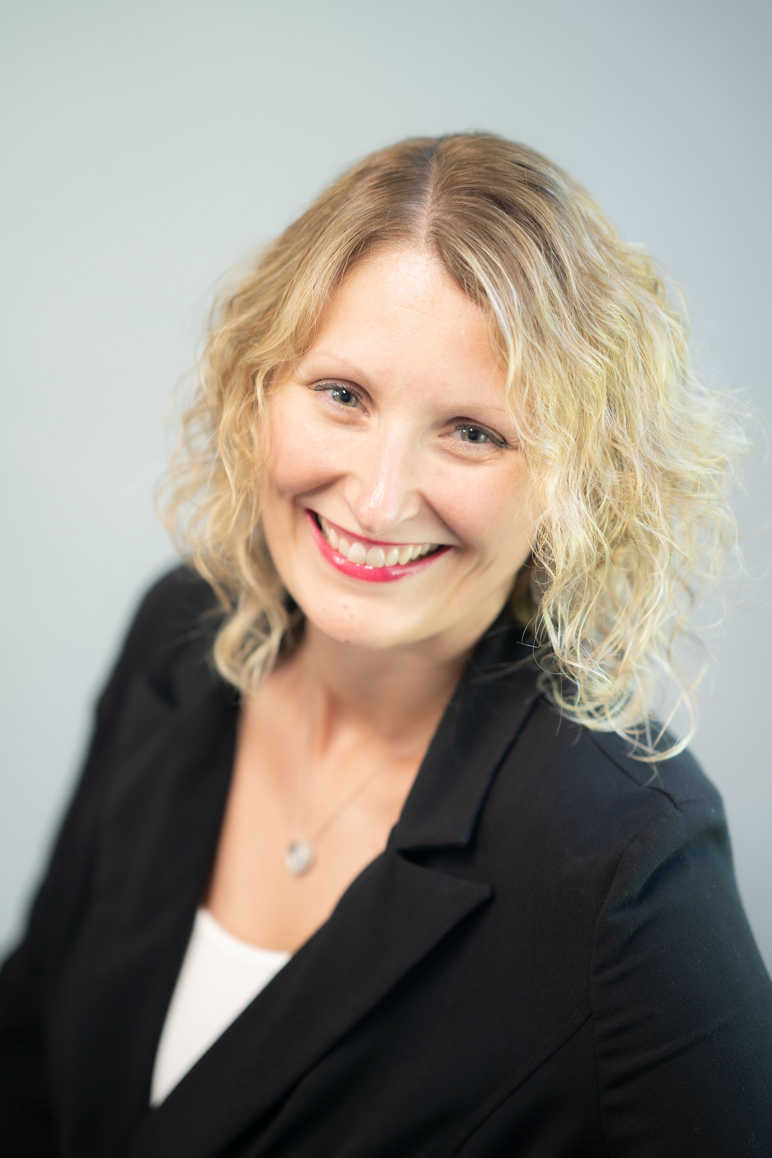 Professional headshot featuring someone with curly blonde hair wearing a black blazer and white top.