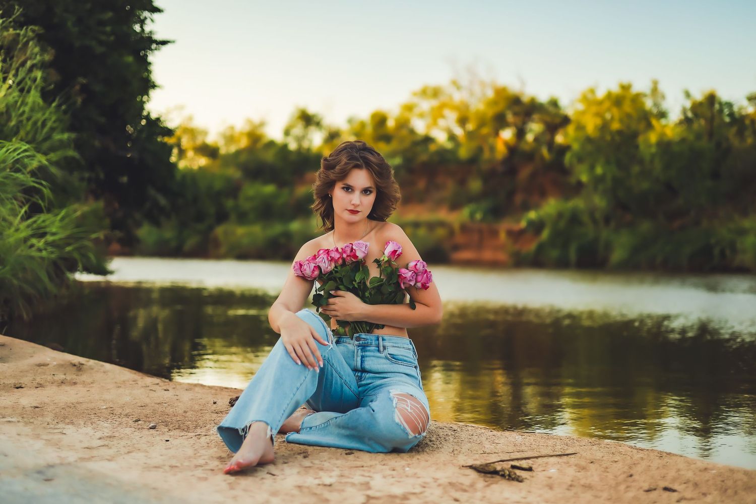 A person in jeans and floral top sits contemplatively by a serene lake at sunset.