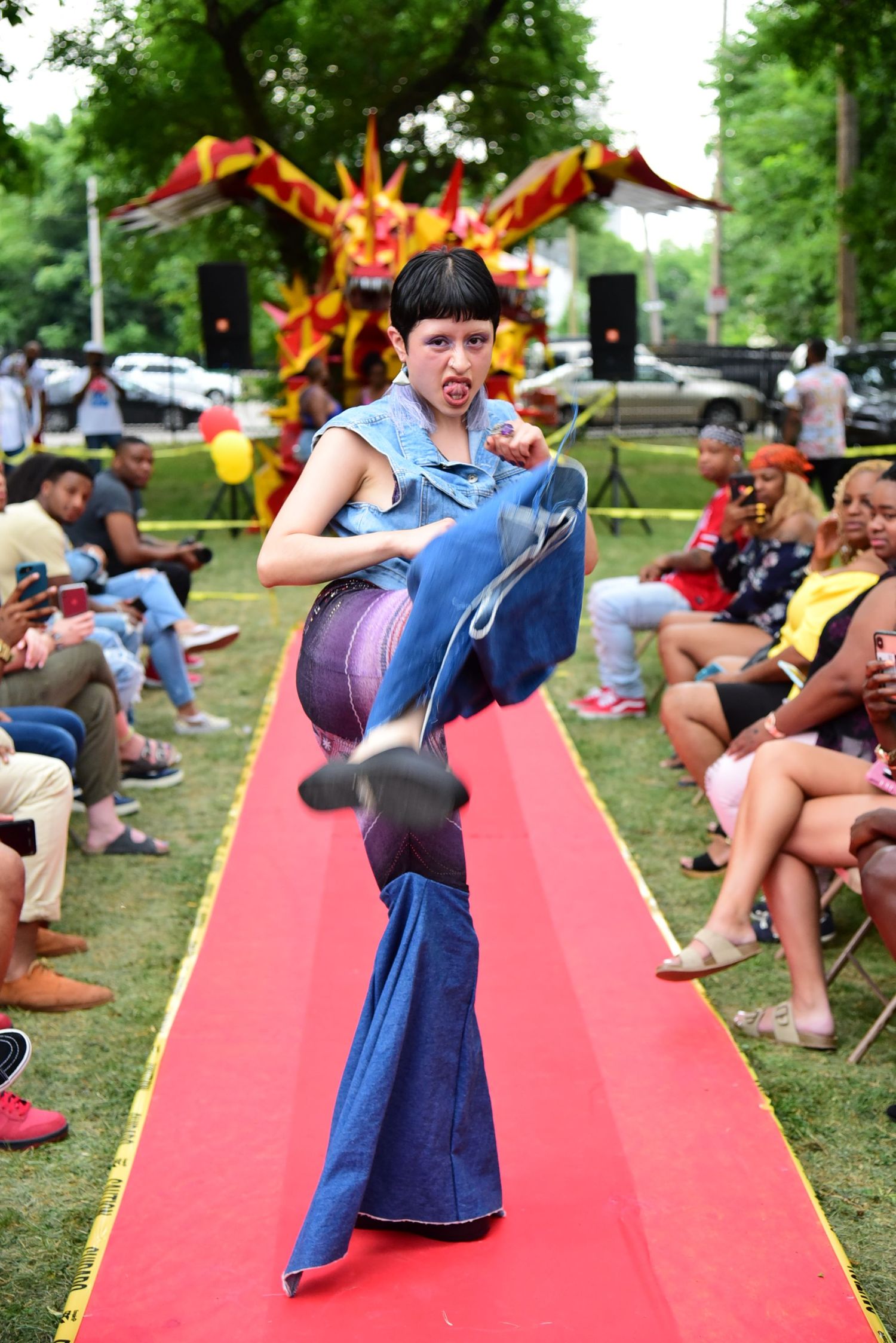 People walk down a red runway at an outdoor fashion show with colorful decorations.