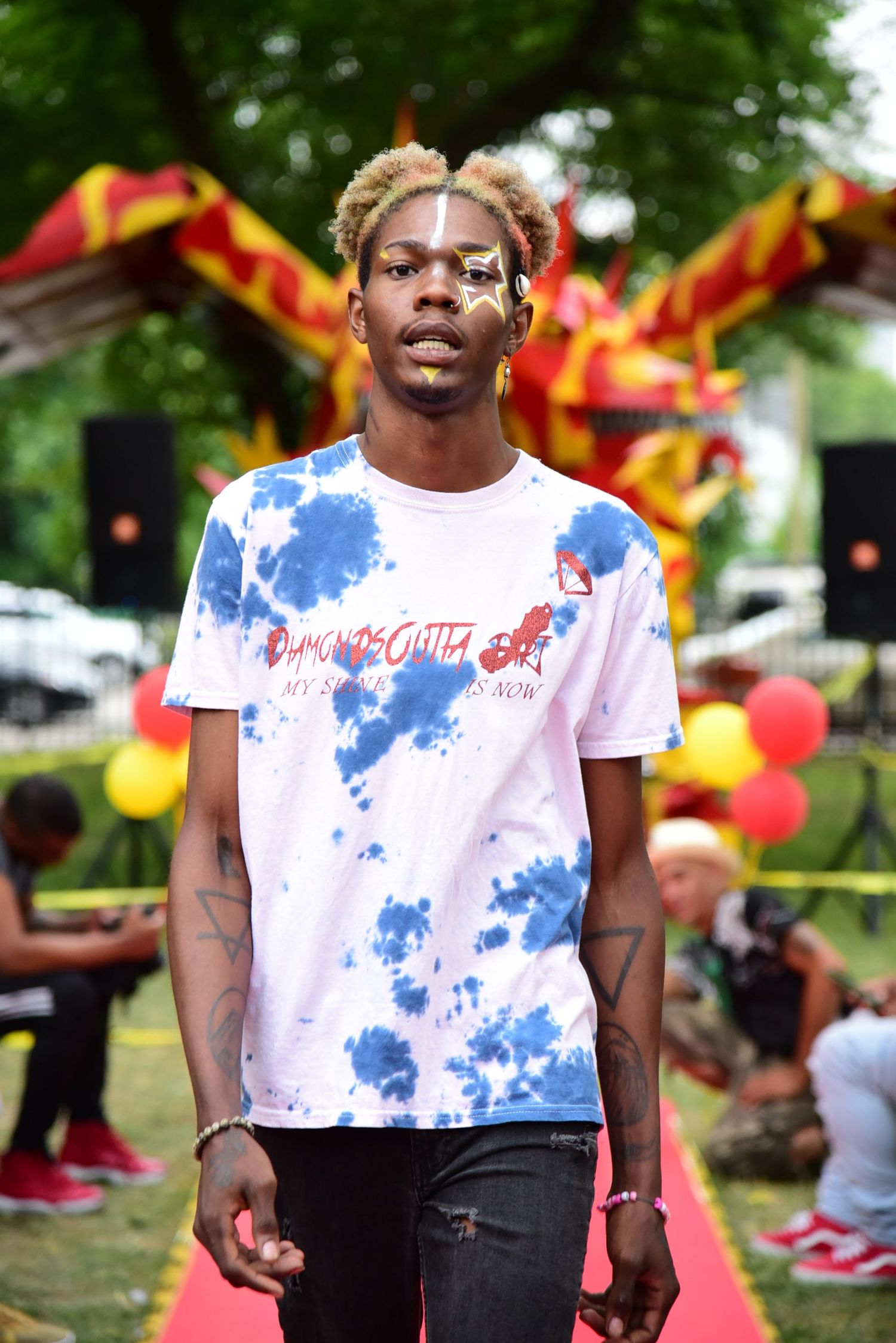 Person wearing a tie-dye white and blue t-shirt poses on red carpet at outdoor event.