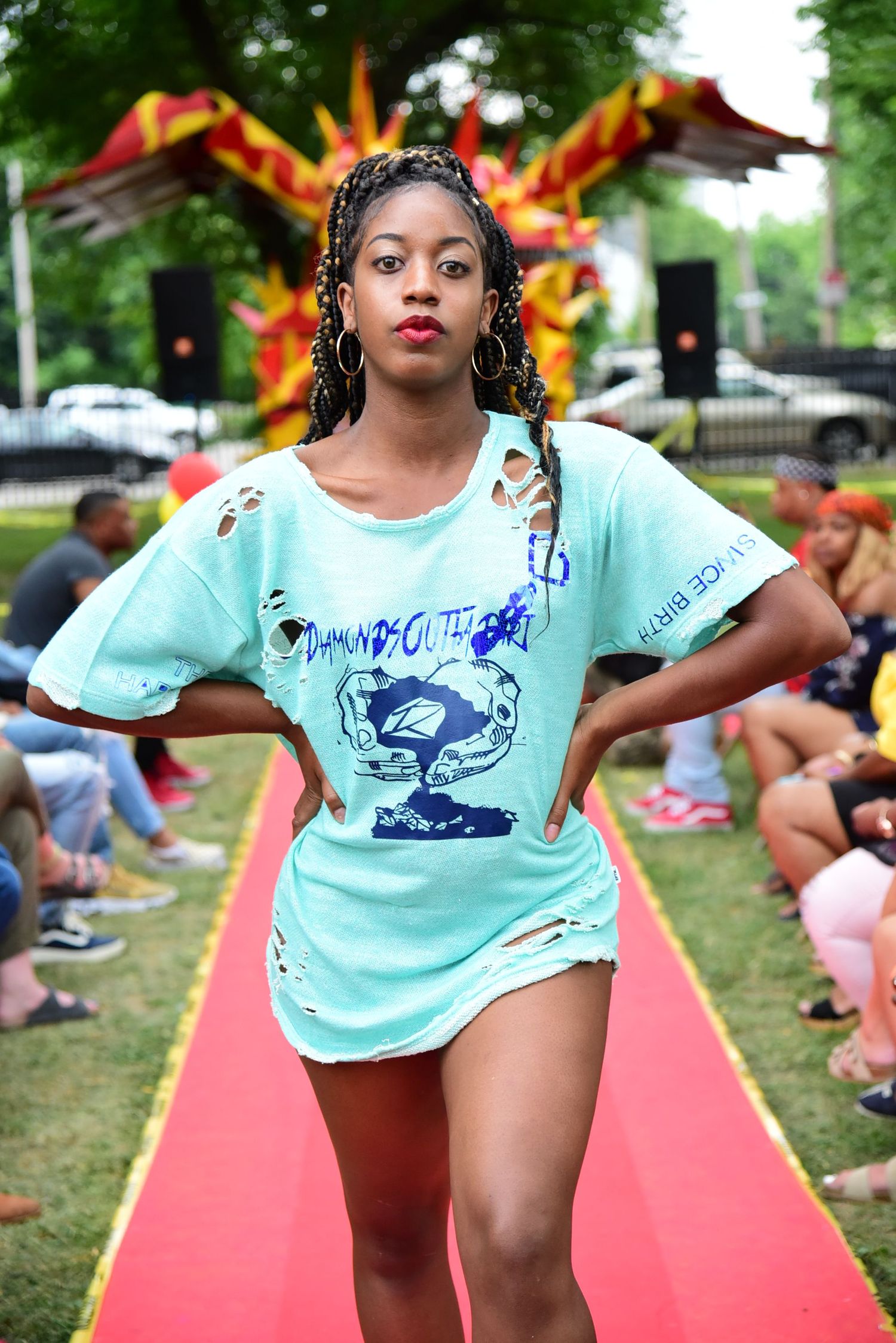 Model in mint green t-shirt walks confidently down red runway at outdoor fashion show.