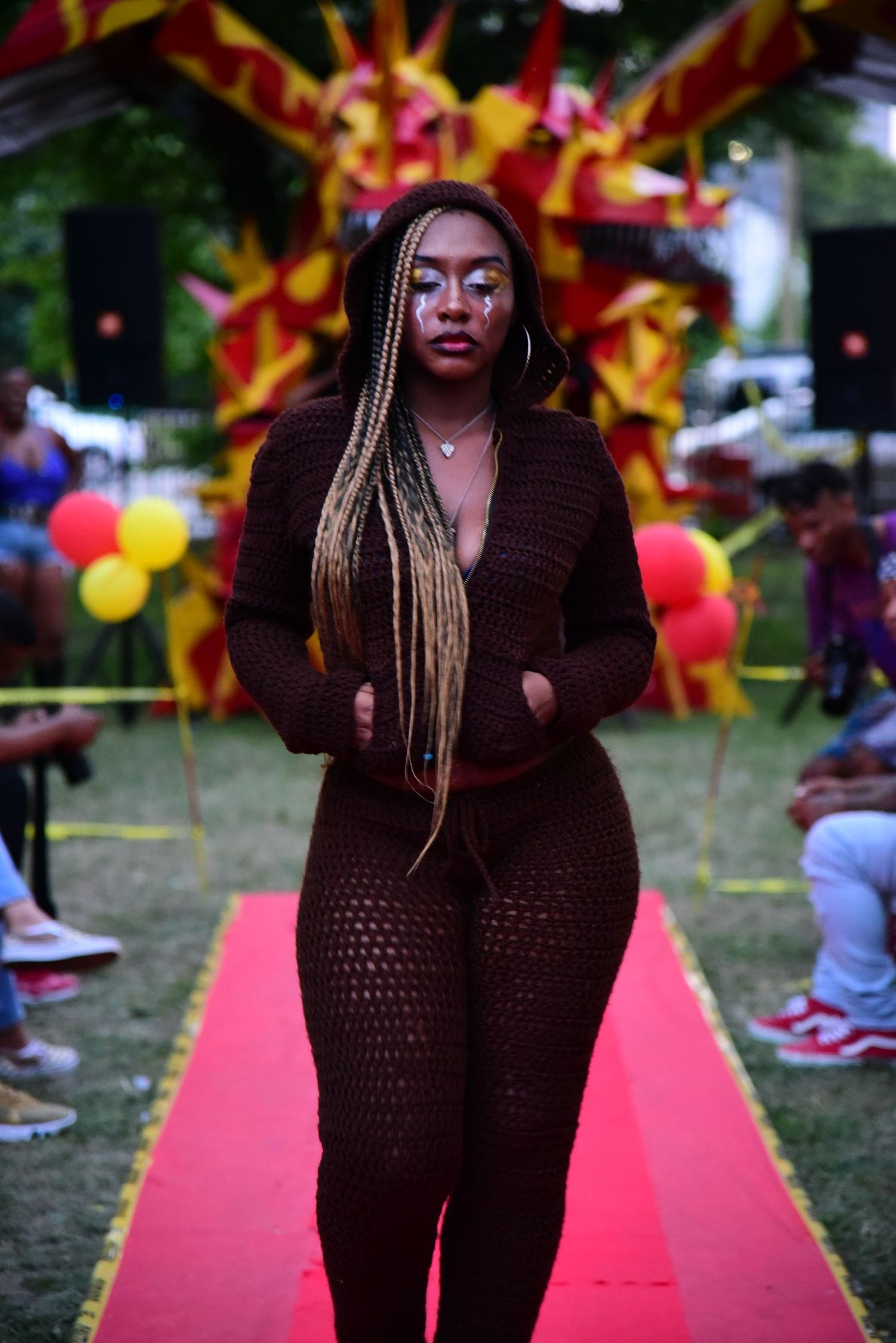 Model walks runway in brown crochet outfit at outdoor fashion show with balloon decorations.