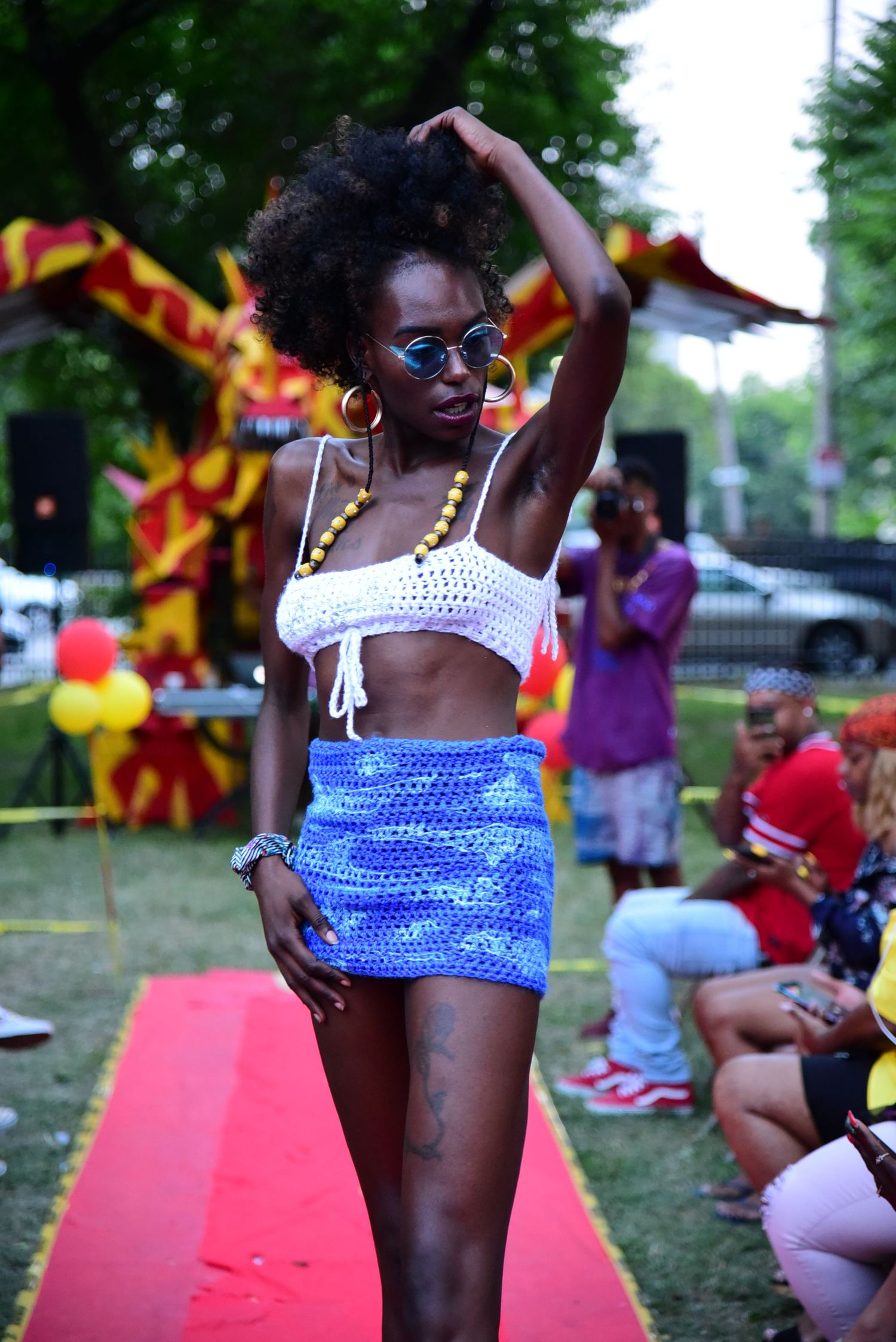 Model in blue crochet top poses on red carpet at outdoor summer fashion event.